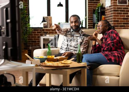 African American romantic partners sitting close together on couch, eating fast food, talking and using remote to browse television. Boyfriend pointing at screen as girlfriend switches tv channels. Stock Photo