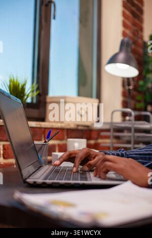 Closeup of black professional scrolling on laptop touchpad in modern brick wall office, updating project slides. African American individual typing on keyboard and working on digital device at desk. Stock Photo