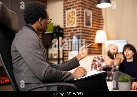 African American counselor takes notes on clipboard, supporting young couple during marriage counseling. Therapist in glasses observes as husband and wife share struggles about love and stress. Stock Photo