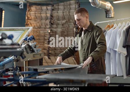 Young Man Operating Screen Printing Machine in Clothing Workshop Stock Photo