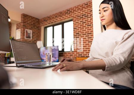 Asian IT specialist works remotely from stylish brick wall apartment, programming across multiple screens. Female tech entrepreneur reviews code and ensures cybersecurity from home. Stock Photo