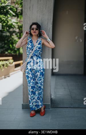 Woman in blue floral jumpsuit leans against a pillar in a sunny urban setting Stock Photo