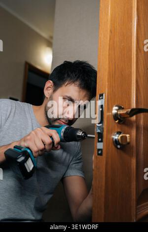Focused bearded young carpenter male installing new door lock using electric screwdriver, engaged in home improvement project, working on diy task Stock Photo