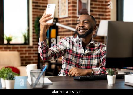 Cheerful remote male employee taking selfie on smartphone during break time at home office desk. Smiling businessman raising hand with mobile device, capturing self portrait picture for social media. Stock Photo