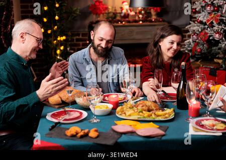 Senior and young people on christmas eve gathering at table to eat homemade food and drink wine, being festive together. Diverse family and friends having fun celebrating december holiday. Stock Photo