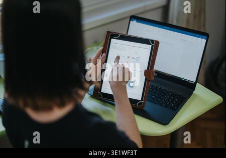 Woman using a tablet and laptop at a home office desk, working on digital charts and presentations Stock Photo