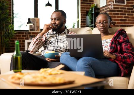 Black woman handling emails on laptop while bearded partner relaxing with snacks. African american spouses on couch, relaxing with food and entertainment in comfortable living room. Stock Photo