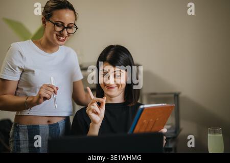 Two women collaborating on a laptop and tablet in a casual home office setting, smiling and discussing work Stock Photo