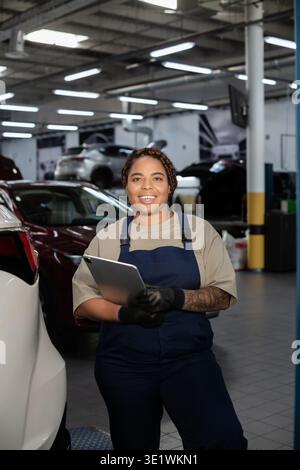 Young African American woman in overalls confidently checks car details in a garage. Stock Photo