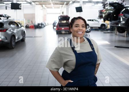 Young African American woman in overalls confidently works among cars in a vibrant garage. Stock Photo