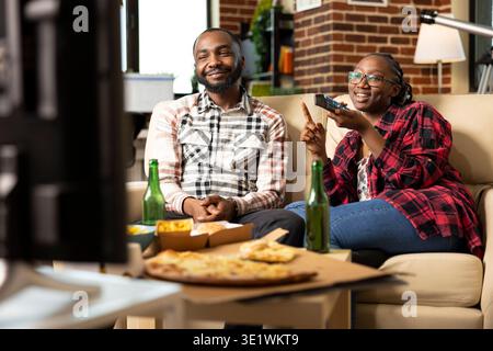 Smiling black man sitting on sofa beside girlfriend with remote, watching movie at home. Young couple relaxing with digital entertainment and takeout food during weekend in brick wall apartment. Stock Photo