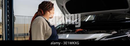 Young African American woman in overalls inspects a car engine in a bustling garage, banner Stock Photo