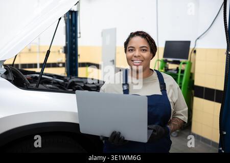 Female mechanic with a laptop smiles while inspecting a car in an auto garage workshop Stock Photo