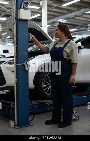 Dedicated female mechanic in overalls confidently troubleshooting a car in a vibrant garage. Stock Photo
