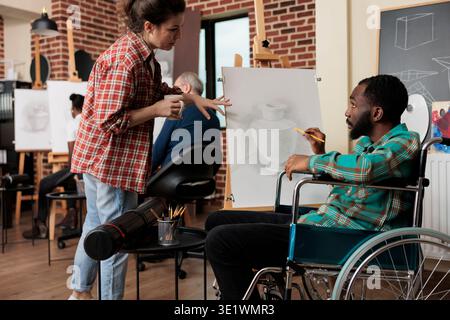 African American disabled guy talking with teacher during group drawing class, improving social skills through collaborative artwork. Teaching art to people with disabilities Stock Photo