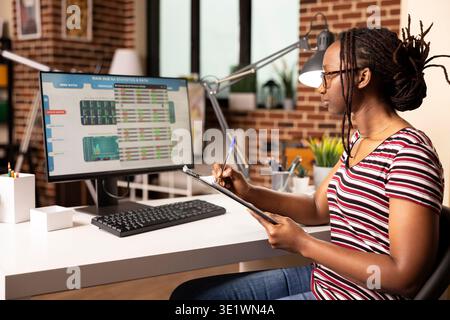 Self employed woman analyzing business charts on desktop pc, comparing metrics with notes from clipboard. Black female manager reviewing financial graphs on computer screen at home. Stock Photo