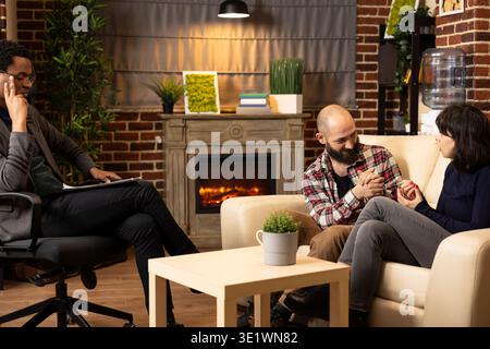 Focused psychotherapist observes tender moment between caucasian woman and man having open conversation about relationship in therapy. Young couple talking as psychiatrist takes notes during counseling Stock Photo