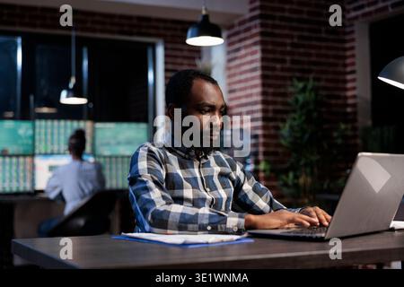 African american business man sitting at desk in office workspace while using laptop to analyze market trend. Professional trader doing technical analysis regarding forex stock trading at night. Stock Photo