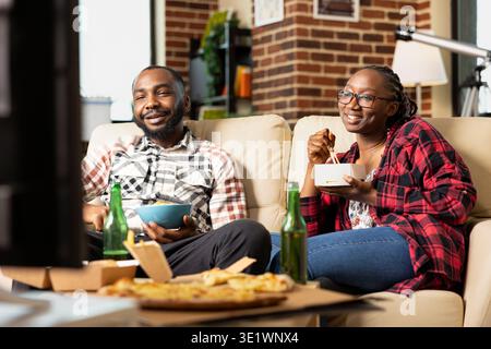 Bearded man and woman with glasses enjoying funny movie together at brick wall apartment. Relaxed weekend moment with snacks in hand, bonding in warm living room on comfortable sofa. Stock Photo