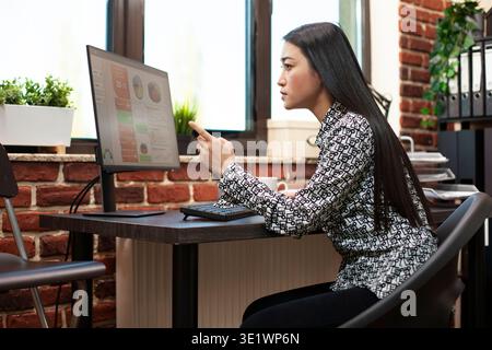 Female analyst pointing at computer screen, closely reviewing financial graphs in startup office. Young businesswoman concentrating on her desktop monitor, looking at company research information. Stock Photo