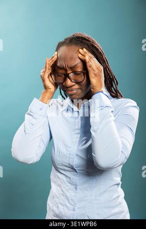 Black female individual feeling sick, standing with eyes shut, pressing her temples in visible discomfort. Exhausted african american woman touching her head, showing signs of a painful migraine. Stock Photo