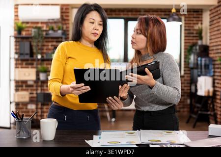 Two female colleagues standing and sharing their strategy ideas in brick wall office. Businesswoman holding a clipboard, comparing project notes with her coworker using a tablet in modern workspace. Stock Photo