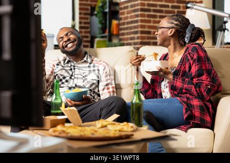 Cheerful black couple enjoying cozy movie hangout at home, laughing while watching TV. African american spouses having relaxed weekend with beer bottles, crisps and takeout food in warm living room. Stock Photo