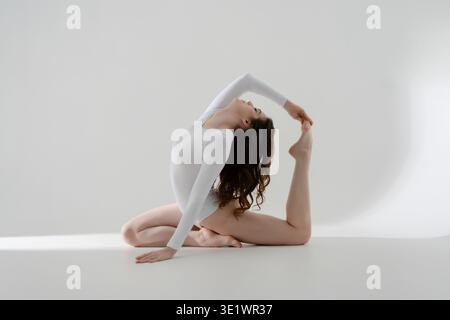 Flexible woman in a white leotard practices yoga, performing a graceful backbend in a minimalist studio. Stock Photo