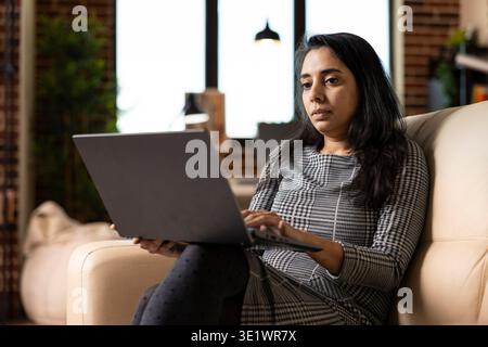 Modern Indian woman seated on couch, focused on editing blog post and researching business trends. Female entrepreneur working remotely with laptop, reviews marketing plans in cozy home environment. Stock Photo