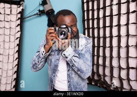 Focused black man using professional photography gear against an isolated background. Portrait of a male photographer standing near softboxes and studio lights, capturing images with his DSLR camera. Stock Photo