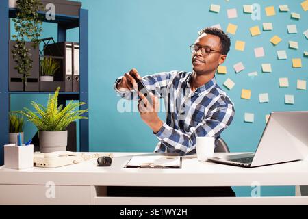 African American male analyst uses cellphone for video games, balancing work and leisure in marketing company. Young businessman seated at desk, enjoying a game on his mobile device. Stock Photo