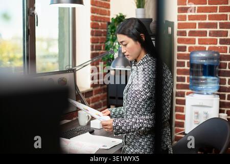 Professional asian woman holds papers in her hands, standing to assess research documents at her office desk. Female entrepreneur reviewing financial paperwork for company projects, near the window. Stock Photo