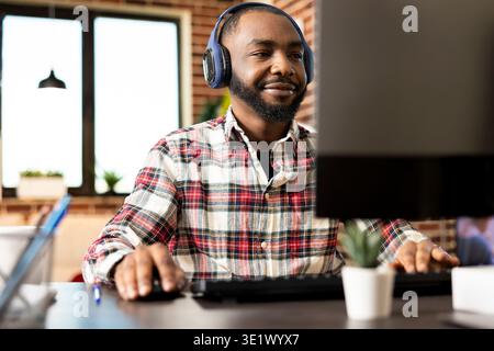 Male financial analyst wearing wireless headset works remotely from home office, using mouse and keyboard at desk. Bearded black man reviewing business reports on desktop pc in brick wall room. Stock Photo