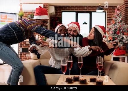 Cheerful coworkers fighting for game console joystick while celebrating christmas in decorated office. Happy diverse colleagues having video gaming competition at new year corporate party Stock Photo