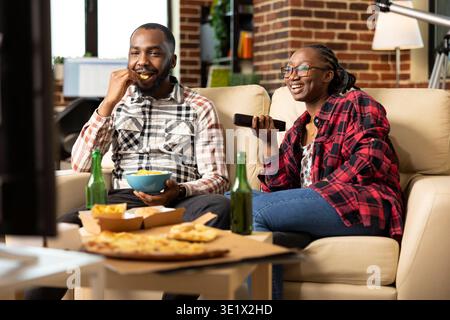 Portrait of cheerful romantic partners watching movies at home and eating snacks and pizza, sitting on couch together. Black man munching on crisps as woman increases tv volume using remote control. Stock Photo