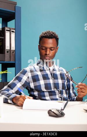 Portrait of a black man holding his spectacles and reviewing paperwork at his desk. African american entrepreneur with glasses and a pen in hand, seated with a clipboard on table, reading documents. Stock Photo