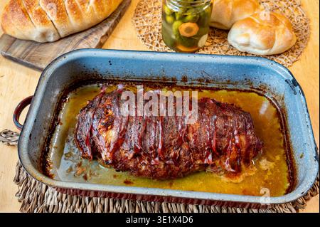 Baked minced meat loaf with bacon in old pan, fresh bun and pickled cucumber on table. Stock Photo