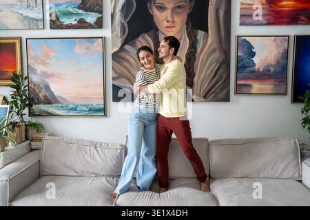 Happy married couple of painters man and woman hugging on sofa in front of home gallery exhibition. Stock Photo