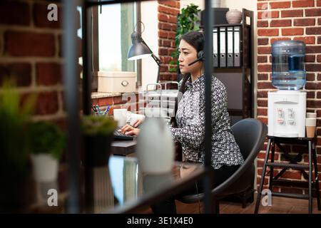 Female call center agent wearing a headset and working on her computer in a well organized office with brick walls and water dispenser nearby. Asian woman with wired headphones, using her desktop pc. Stock Photo