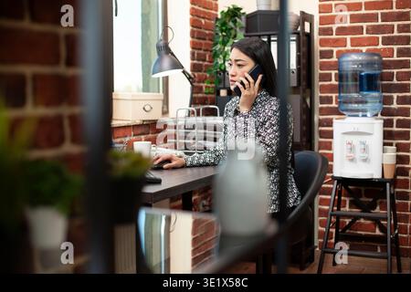 Female entrepreneur using her smartphone for a client conversation and looking at her desktop monitor. Asian manager talks to her colleague on her mobile device referencing updates from her computer. Stock Photo