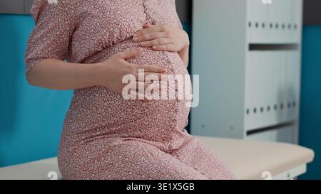 Pregnant patient attending checkup visit with obstetrician, sitting on bed in medical office. Woman with pregnancy belly preparing for childbirth, waiting on health specialist. Close up. Stock Photo
