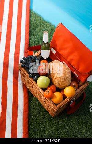 Sunlit picnic basket filled with fresh fruit and bread. Bottle of wine sits beside the basket. Orange and red striped towel lies beside the basket. Gr Stock Photo