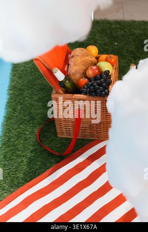 Wicker picnic basket filled with fresh fruit and bread. Red strap drapes casually over grassy ground. Orange and white striped blanket lies below. Whi Stock Photo