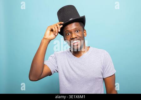 Smiling young african american man in a costume prop preparing for a show. Cheerful black magician in a casual tee, getting ready for a classic magic show while adjusting his top hat. Stock Photo
