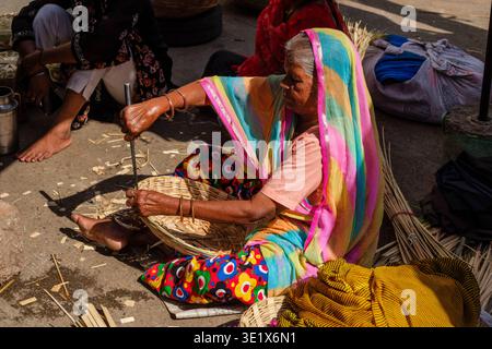 An elderly woman in a colourful sari sits on the ground in Udaipur, using a metal tool to meticulously finish the rim of a hand-woven basket. Udaipur Stock Photo