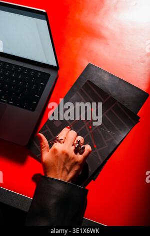 Hand rests on dark map atop vibrant red surface. Laptop sits beside map, suggesting planning or navigation. Fingered hand wears rings, adding personal Stock Photo
