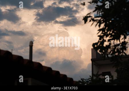 Dramatic clouds glow softly above rooftops. Silhouettes of buildings frame the sky's gentle hue. Branches cast dark shapes against the light. Sunset r Stock Photo