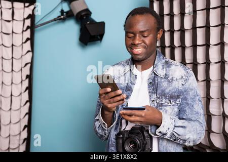 African American man standing in his studio, happily making an online purchase with his cellphone. Cheerful male photographer entering his credit card details on his mobile, completing a transaction. Stock Photo