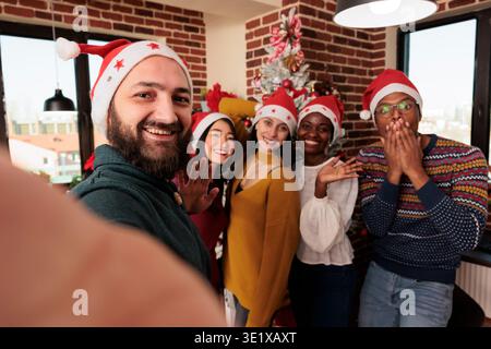 Smiling man in santa hat taking slefie with colleagues in decorated office at christmas corporate party. Coworkers looking at camera while posing for mobile phone group photo at festive workplace Stock Photo