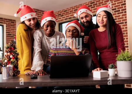 Cheerful colleagues checking sales annual report results on laptop before celebrating christmas in office. Smiling happy coworkers working for project deadline together in festive decorated workplace Stock Photo